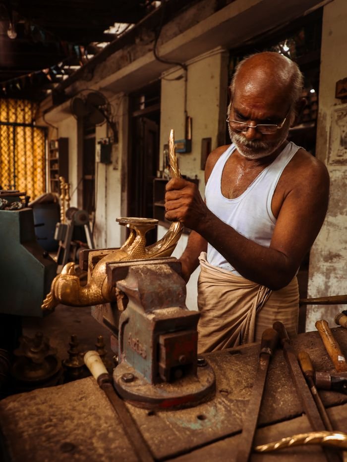 Elderly artisan crafting metal sculpture in Natchiarkoil, Tamil Nadu workshop indoors.