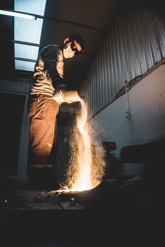 A worker in safety gear pours molten metal, illuminating the factory space with fiery light.